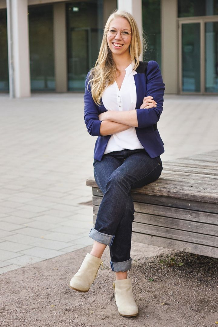Eine Person mit langen Haaren sitzt auf einer Bank im Freien. Sie trägt eine Brille, einen blauen Blazer, ein weißes Hemd, dunkelblaue Jeans und helle Stiefeletten. Die Beine sind übereinandergeschlagen. Im Hintergrund ist ein Gebäude.