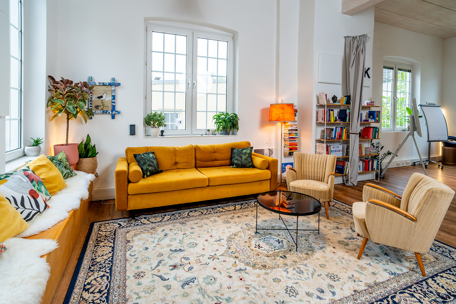 Helles Wohnzimmer mit gelbem Sofa, zwei beige Sesseln und rundem Glastisch auf einem gemusterten Teppich. An der Wand stehen Bücherregale, eine orangefarbene Stehlampe und mehrere Pflanzen auf Fensterbänken. Holzfußboden und weiße Wände.