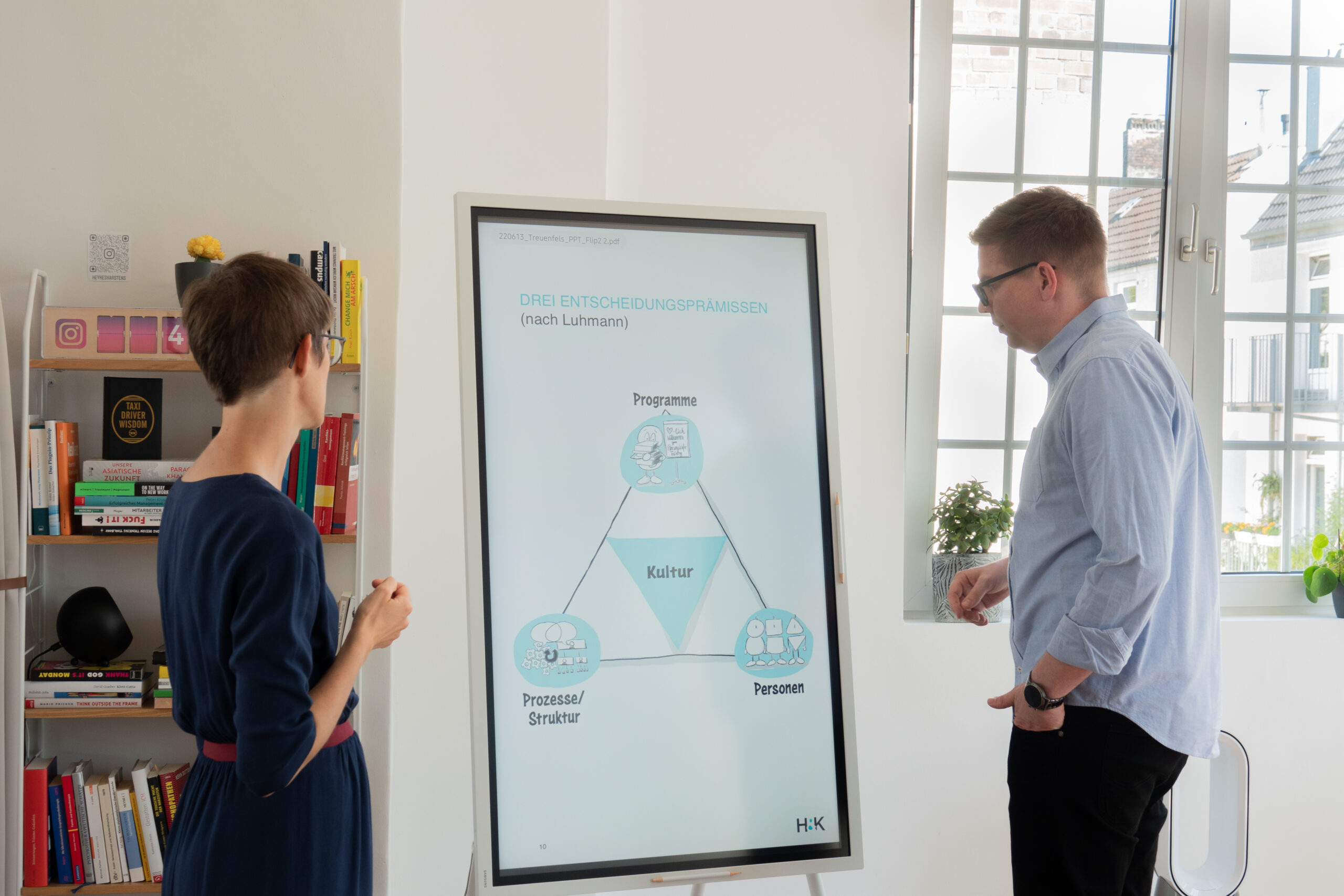 Eine Frau und ein Mann stehen vor einem vertikalen Bildschirm, der ein Diagramm mit den Begriffen Programme, Kultur, Personen und Prozesse/Struktur zeigt. Im Hintergrund sind ein Bücherregal und Fenster zu sehen.