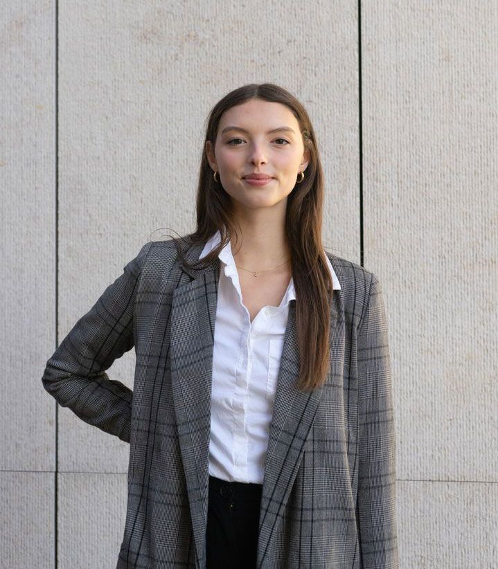 Eine Person in einem karierten Blazer und weißem Hemd steht vor einer hellen, strukturierten Wand. Die Person hat lange, offene Haare und hält eine Hand in die Hüfte gestützt.