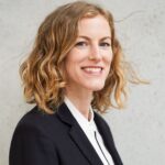 Eine lächelnde Person mit lockigem, schulterlangem Haar steht vor einer grauen Wand. Sie trägt einen schwarzen Blazer und ein weißes Hemd.