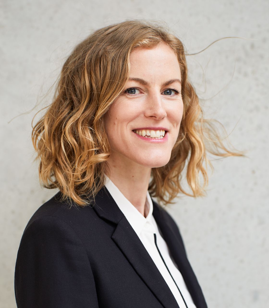 Eine lächelnde Person mit lockigem, schulterlangem Haar steht vor einer grauen Wand. Sie trägt einen schwarzen Blazer und ein weißes Hemd.