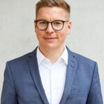 Eine Person mit Brille, hellem Haar und einem blauen Anzug trägt ein weißes Hemd. Der Hintergrund ist eine schlichte, helle Wand. Die Person blickt freundlich in die Kamera.