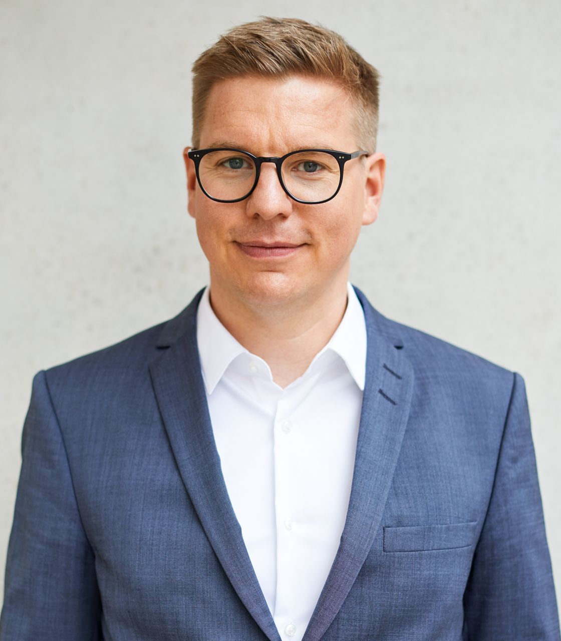 Eine Person mit Brille, hellem Haar und einem blauen Anzug trägt ein weißes Hemd. Der Hintergrund ist eine schlichte, helle Wand. Die Person blickt freundlich in die Kamera.