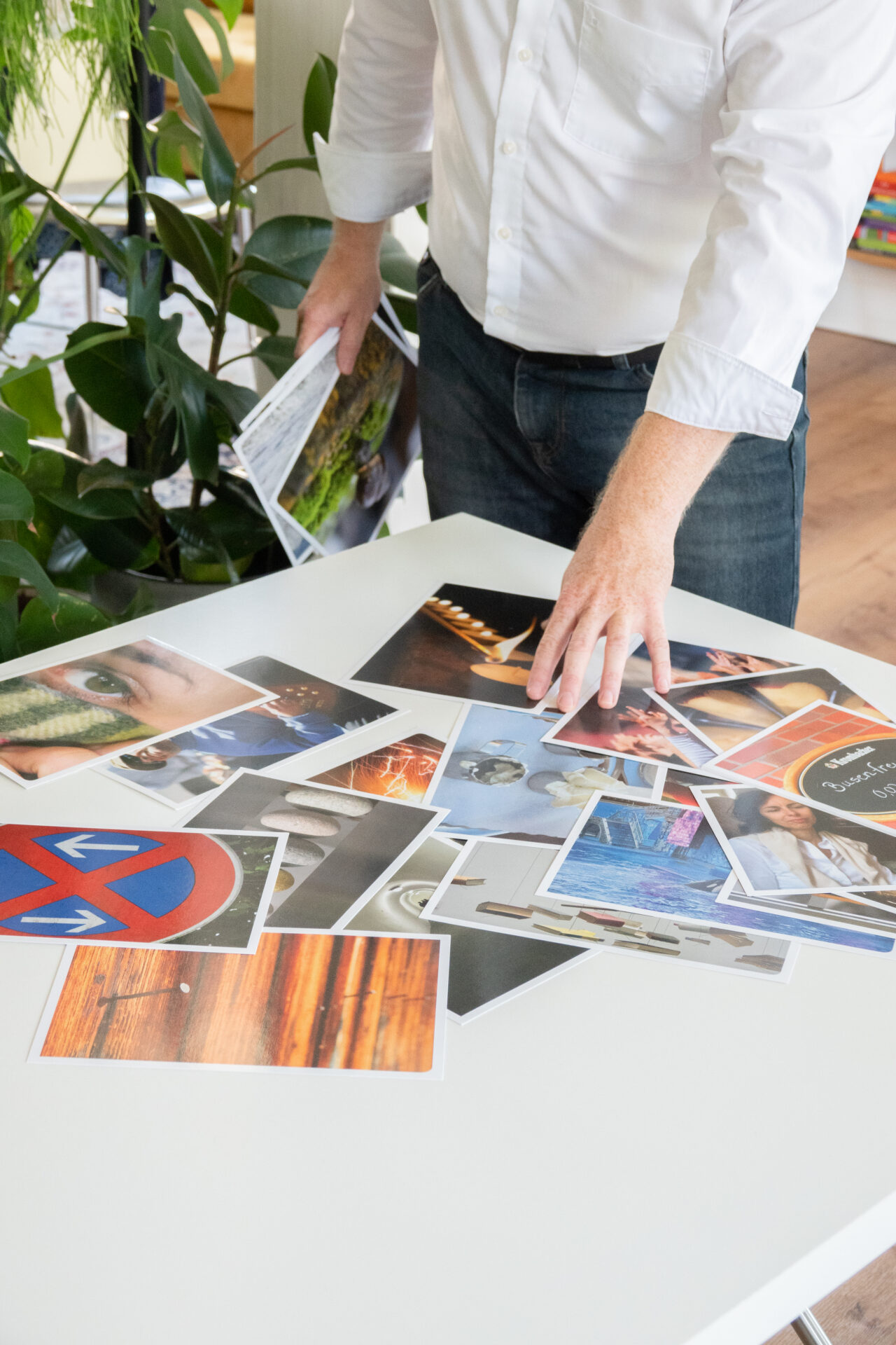 Eine Person in einem weißen Hemd steht neben einem Tisch und sortiert mehrere Fotos, die auf dem Tisch verstreut liegen. Im Hintergrund sind grüne Pflanzen sichtbar.