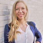 Eine lächelnde Person mit langen blonden Haaren und Brille steht vor einer hellen Ziegelwand. Sie trägt ein weißes Hemd und ein blaues Sakko. Die Arme sind entspannt an der Seite.