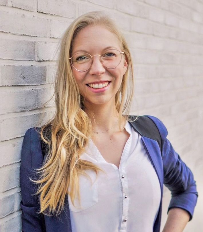 Eine lächelnde Person mit langen blonden Haaren und Brille steht vor einer hellen Ziegelwand. Sie trägt ein weißes Hemd und ein blaues Sakko. Die Arme sind entspannt an der Seite.
