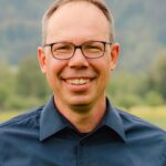 Eine Person mit Brille und einem dunklen Hemd lächelt in die Kamera. Der Hintergrund ist unscharf und zeigt eine grüne, natürliche Landschaft.