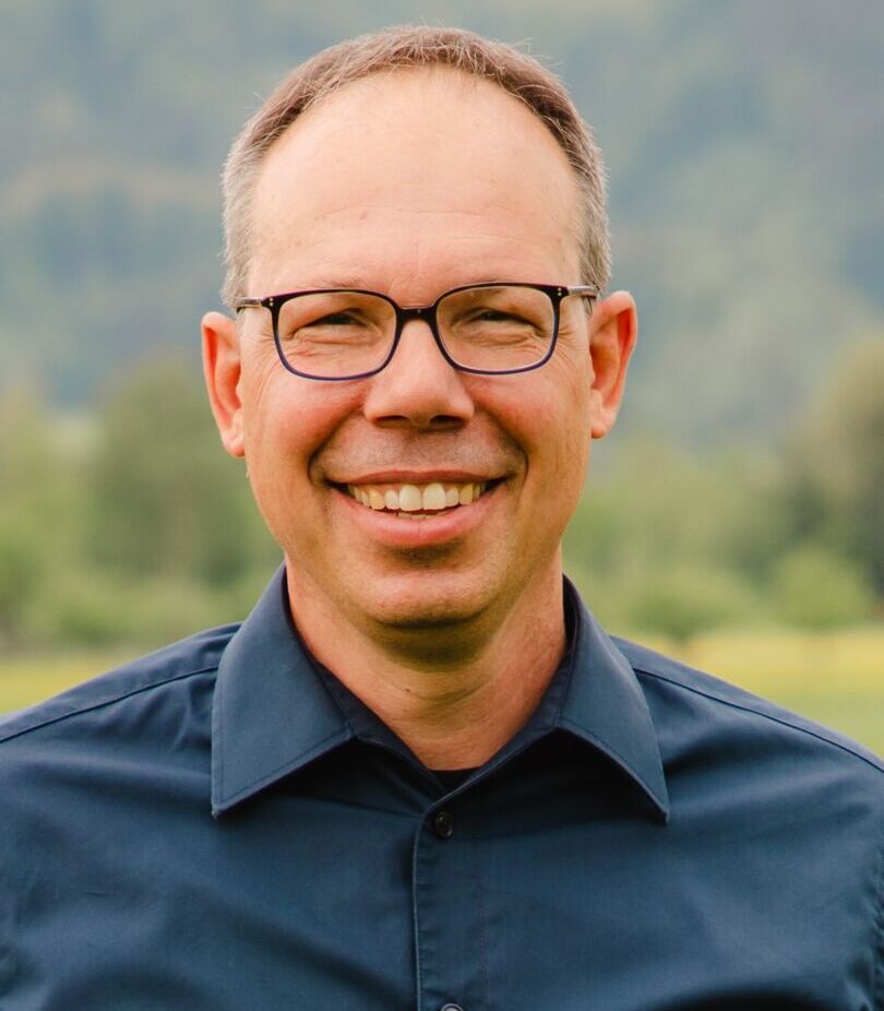 Eine Person mit Brille und einem dunklen Hemd lächelt in die Kamera. Der Hintergrund ist unscharf und zeigt eine grüne, natürliche Landschaft.