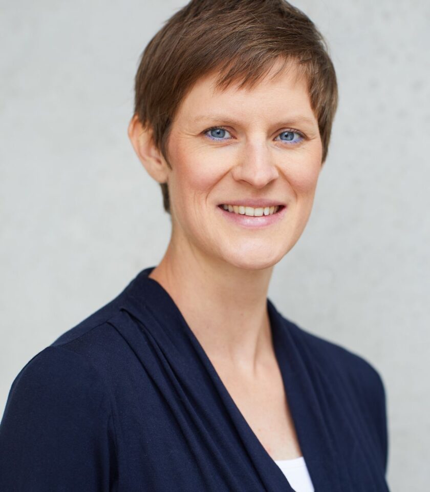 Porträt einer lächelnden Person mit kurzem, braunem Haar, die einen dunkelblauen Blazer und ein weißes Oberteil trägt. Der Hintergrund ist neutral und hellgrau.