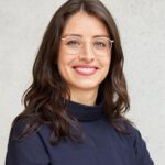 Eine Person mit langen, dunklen Haaren und Brille lächelt in die Kamera. Sie trägt ein dunkelblaues Oberteil. Der Hintergrund ist neutral und hell.