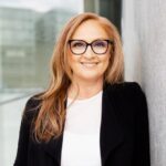 Eine Person mit langen, braunen Haaren trägt eine Brille, ein weißes Oberteil und ein schwarzes Jackett. Sie lehnt an einer grauen Wand und lächelt. Im Hintergrund sind moderne Glasarchitektur und verschwommene Gebäude sichtbar.