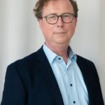 Ein Mann mittleren Alters mit Brille, braunem Haar und hellem Teint trägt ein blaues Hemd und ein dunkles Sakko. Er steht vor einem neutralen, hellen Hintergrund.