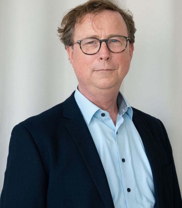 Ein Mann mittleren Alters mit Brille, braunem Haar und hellem Teint trägt ein blaues Hemd und ein dunkles Sakko. Er steht vor einem neutralen, hellen Hintergrund.