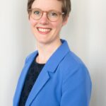 Eine Person mit kurzem Haar und einer Brille lächelt in die Kamera. Sie trägt ein blaues Jackett über einem dunklen Oberteil. Der Hintergrund ist neutral.