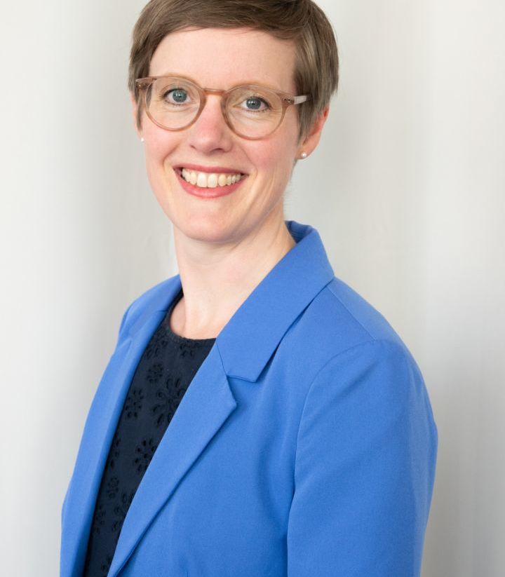 Eine Person mit kurzem Haar und einer Brille lächelt in die Kamera. Sie trägt ein blaues Jackett über einem dunklen Oberteil. Der Hintergrund ist neutral.