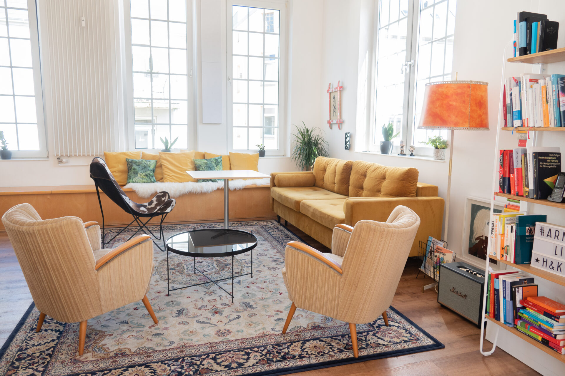 Ein Wohnzimmer mit großen Fenstern. Es gibt ein gelbes Sofa, zwei beige Sessel, einen runden schwarzen Tisch und einen bunten Teppich. Bücherregale und eine orangefarbene Lampe ergänzen die Einrichtung. Pflanzen sind dekorativ verteilt.