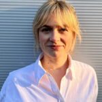 Eine Person mit blondem Haar trägt ein weißes Hemd und steht vor einer geriffelten, grauen Wand. Sie lächelt leicht und blickt in die Kamera. Die Sonne scheint von der Seite und beleuchtet ihr Gesicht.