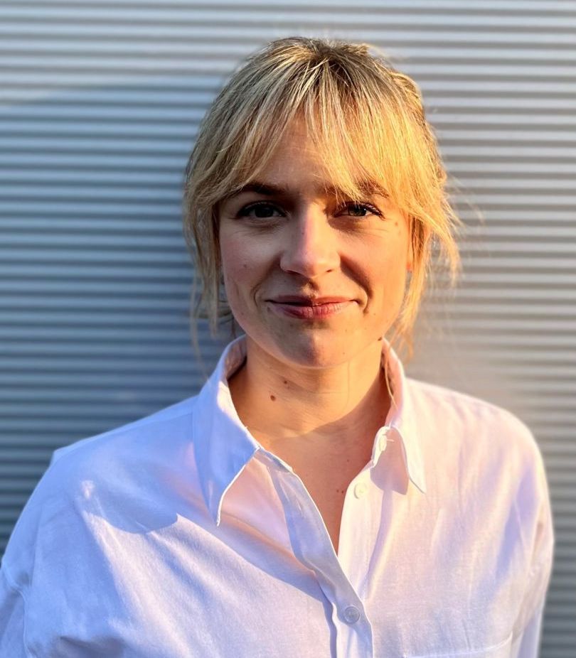 Eine Person mit blondem Haar trägt ein weißes Hemd und steht vor einer geriffelten, grauen Wand. Sie lächelt leicht und blickt in die Kamera. Die Sonne scheint von der Seite und beleuchtet ihr Gesicht.