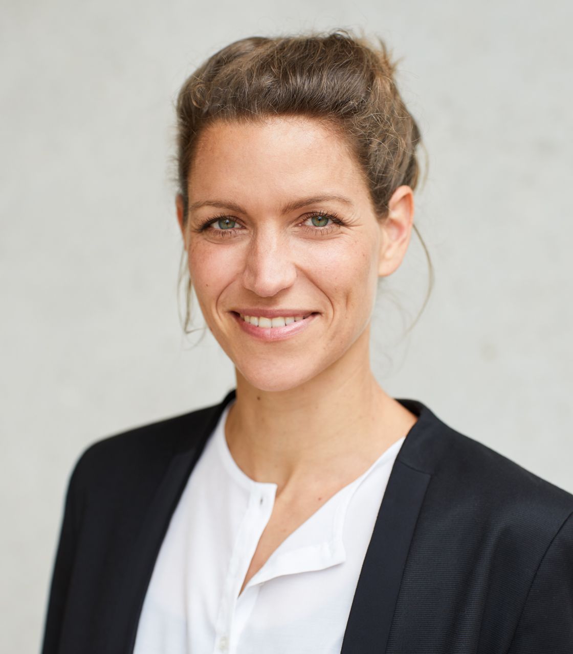 Porträt einer lächelnden Person mit dunklem, geschmeidigem Haarknoten. Sie trägt ein weißes Oberteil und einen dunklen Blazer. Der Hintergrund ist neutral und hell.