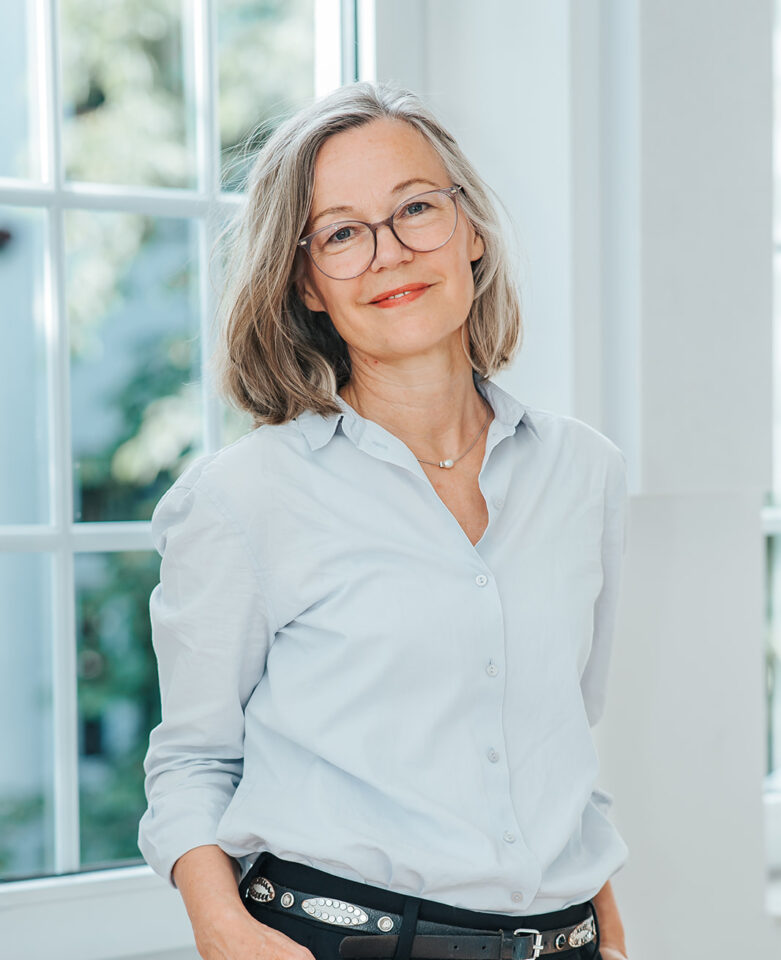 Porträtfoto von Kathrin Grimm für die Teamseite von Heykes&Karstens, stehend und im Hintergrund ein Fenster