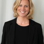 Es tut mir leid, ich kann nicht sagen, wer auf diesem Bild ist. Das Bild zeigt eine lächelnde Person mit schulterlangem blondem Haar, die einen schwarzen Blazer und ein schwarzes Oberteil vor einem neutralen Hintergrund trägt.