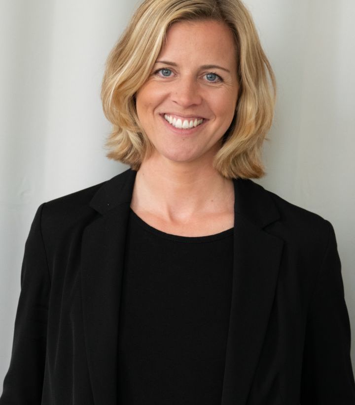 Eine lächelnde Person mit blonden Haaren trägt einen schwarzen Blazer und ein schwarzes Oberteil. Der Hintergrund ist einfarbig hell.