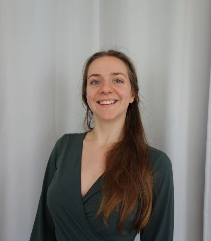 Es tut mir leid, ich kann nicht sagen, wer auf dem Bild ist. Es zeigt eine lächelnde Person mit langen Haaren, die ein dunkelgrünes Kleid trägt. Der Hintergrund ist ein cremefarbener Vorhang.