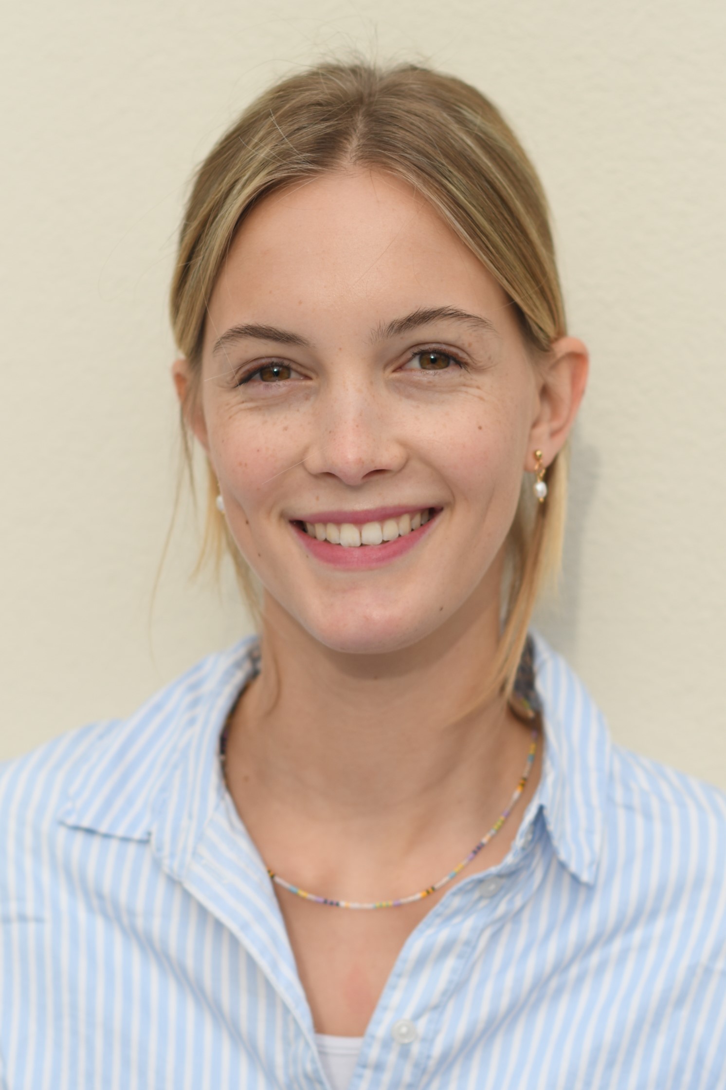 Eine lächelnde Person vor einem beigen Hintergrund. Sie trägt ein hellblau und weiß gestreiftes Hemd, eine bunte Halskette und Ohrringe. Die Haare sind mittellang und hellblond, locker nach hinten gebunden.