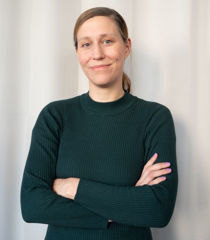 Eine Person mit langem, dunkelblondem Haar, das zu einem Pferdeschwanz gebunden ist, trägt einen dunkelgrünen Pullover. Sie steht mit verschränkten Armen vor einem hellen Hintergrund und lächelt leicht in die Kamera.