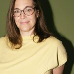 Eine Person mit langen braunen Haaren und Brille trägt ein gelbes T-Shirt und steht vor einem grünen Hintergrund. Sie lächelt leicht.