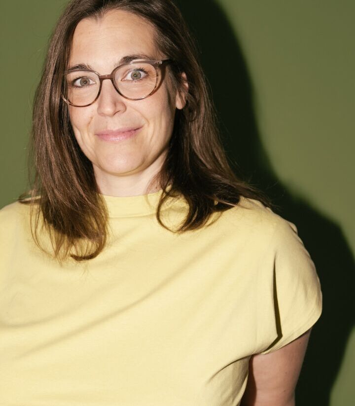 Eine Person mit langen braunen Haaren und Brille trägt ein gelbes T-Shirt und steht vor einem grünen Hintergrund. Sie lächelt leicht.