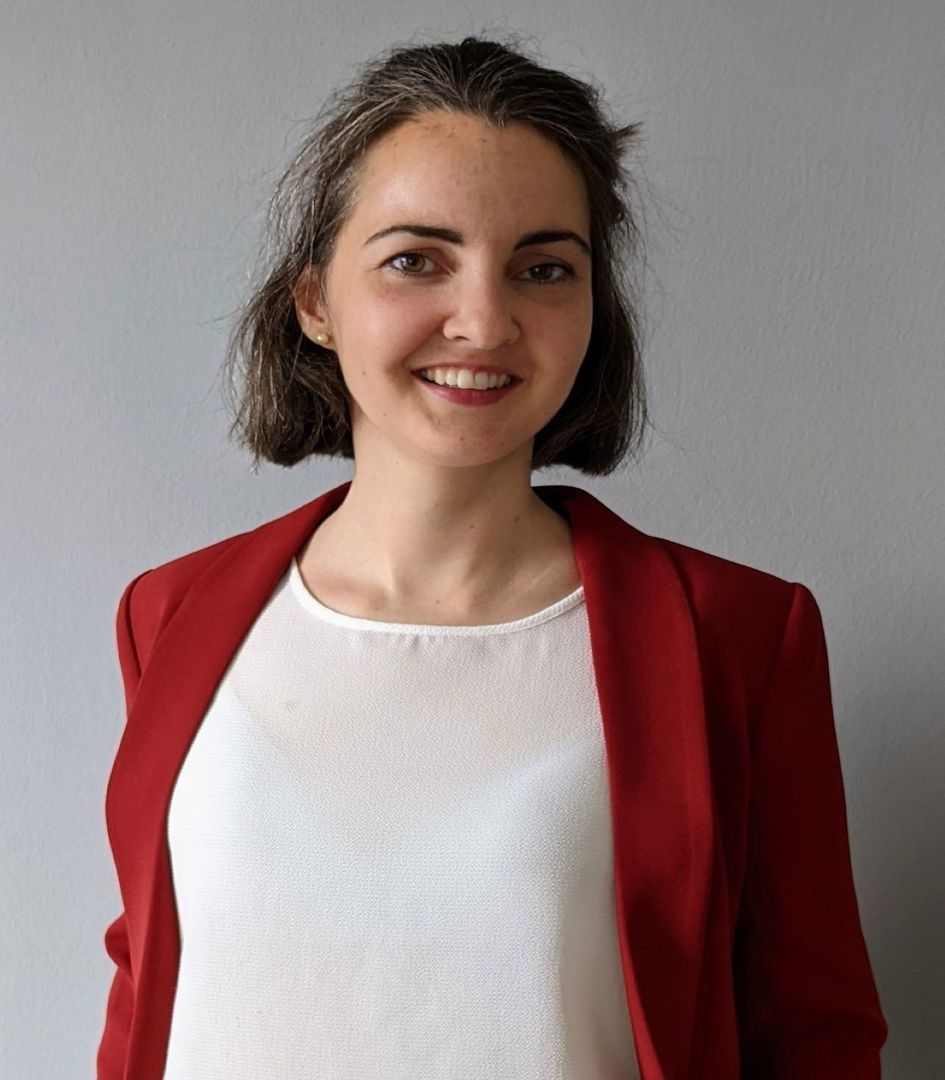 Eine lächelnde Person mit schulterlangem Haar, einem weißen Oberteil und einem roten Blazer steht vor einem neutralen, hellgrauen Hintergrund.