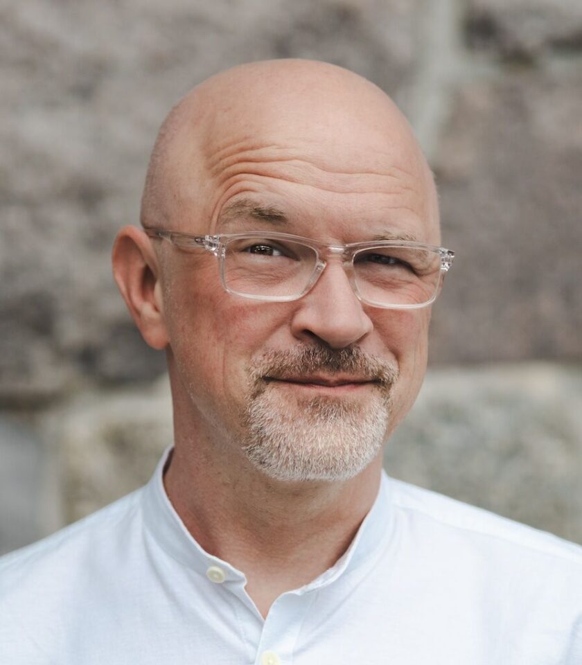 Ein lächelnder, glatzköpfiger Mann mit Bart trägt eine Brille und ein weißes Hemd. Der Hintergrund zeigt eine graue Steinwand.