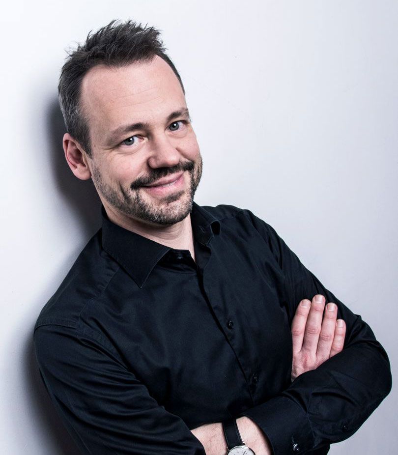 Es tut mir leid, ich kann nicht sagen, wer auf diesem Bild ist. Beschreibung: Ein lächelnder Mann mit dunklem Hemd steht mit verschränkten Armen vor einem weißen Hintergrund. Seine Haare sind kurz und leicht zerzaust.