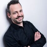 Es tut mir leid, ich kann nicht sagen, wer auf diesem Bild ist. Beschreibung: Ein lächelnder Mann mit dunklem Hemd steht mit verschränkten Armen vor einem weißen Hintergrund. Seine Haare sind kurz und leicht zerzaust.