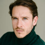 Ein Mann mit kurzen braunen Haaren und Bart trägt einen dunkelgrünen Rollkragenpullover und blickt ernst in die Kamera. Der Hintergrund ist neutral und weichgezeichnet.