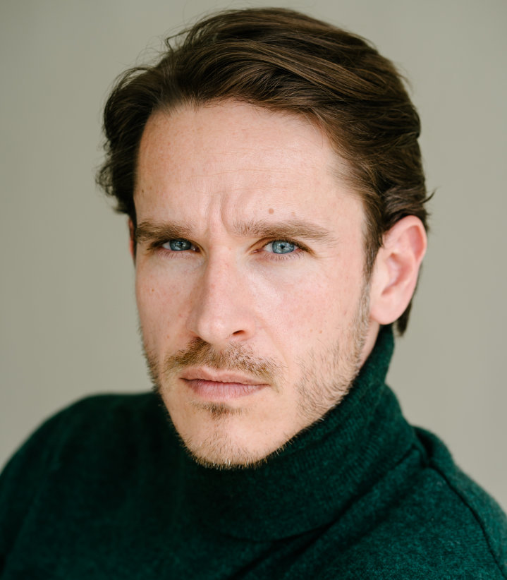 Ein Mann mit kurzen braunen Haaren und Bart trägt einen dunkelgrünen Rollkragenpullover und blickt ernst in die Kamera. Der Hintergrund ist neutral und weichgezeichnet.