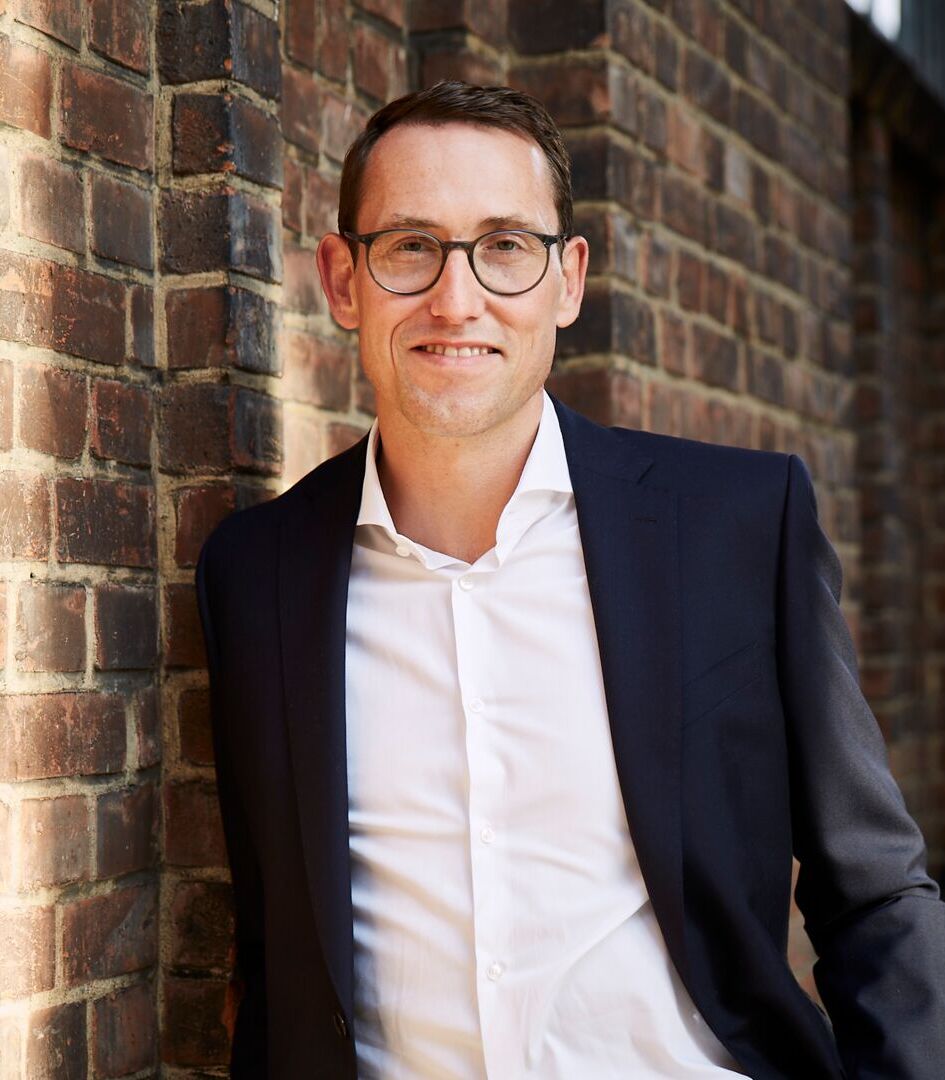 Ein Mann mit Brille trägt ein dunkles Sakko und ein weißes Hemd. Er lehnt lächelnd an einer Backsteinmauer. Das Bild ist tagsüber draußen aufgenommen.