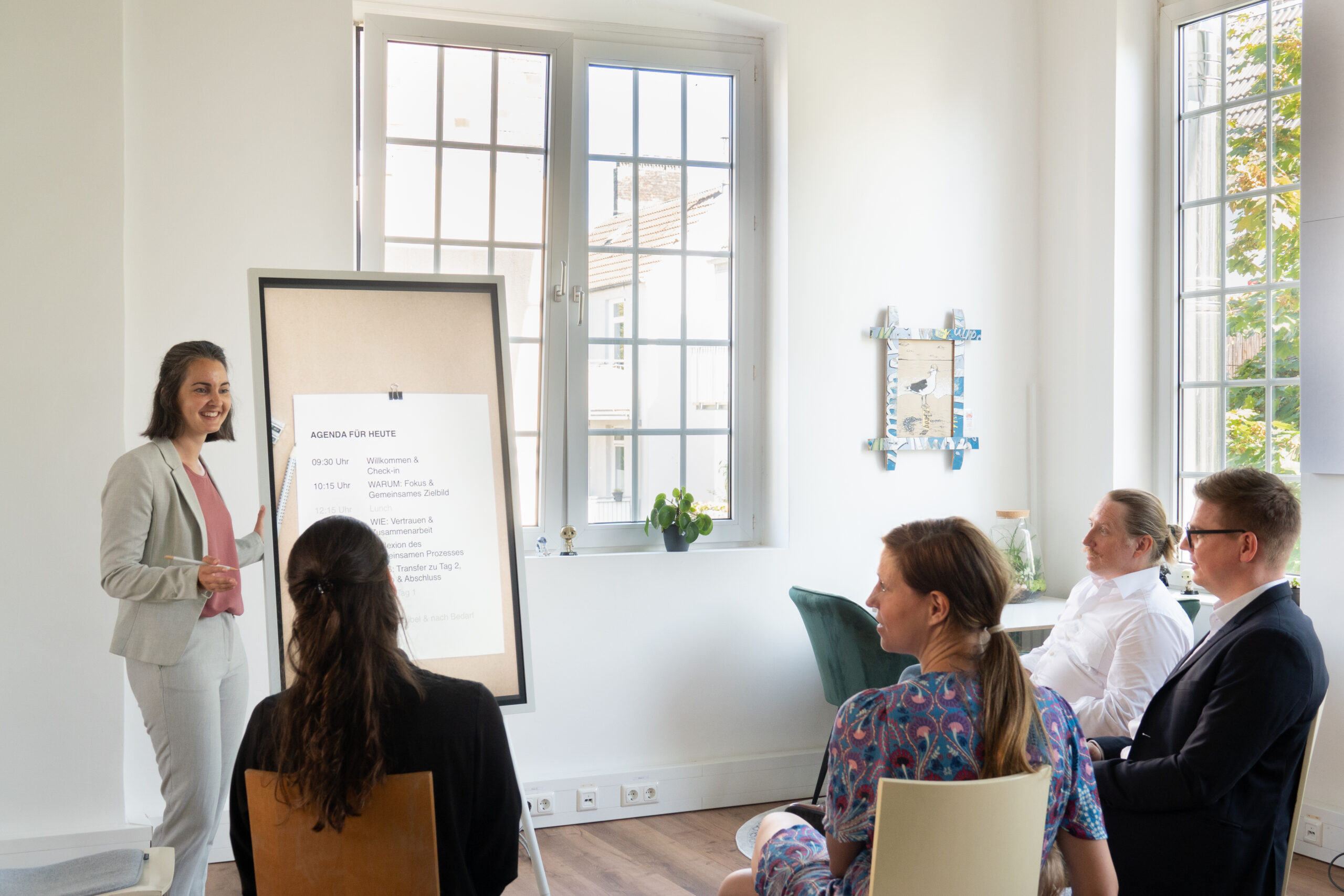 Eine Frau präsentiert in einem hellen Raum vor fünf sitzenden Personen. Sie steht neben einem Flipchart mit Text. Im Hintergrund sind große Fenster und einige Pflanzen zu sehen.
