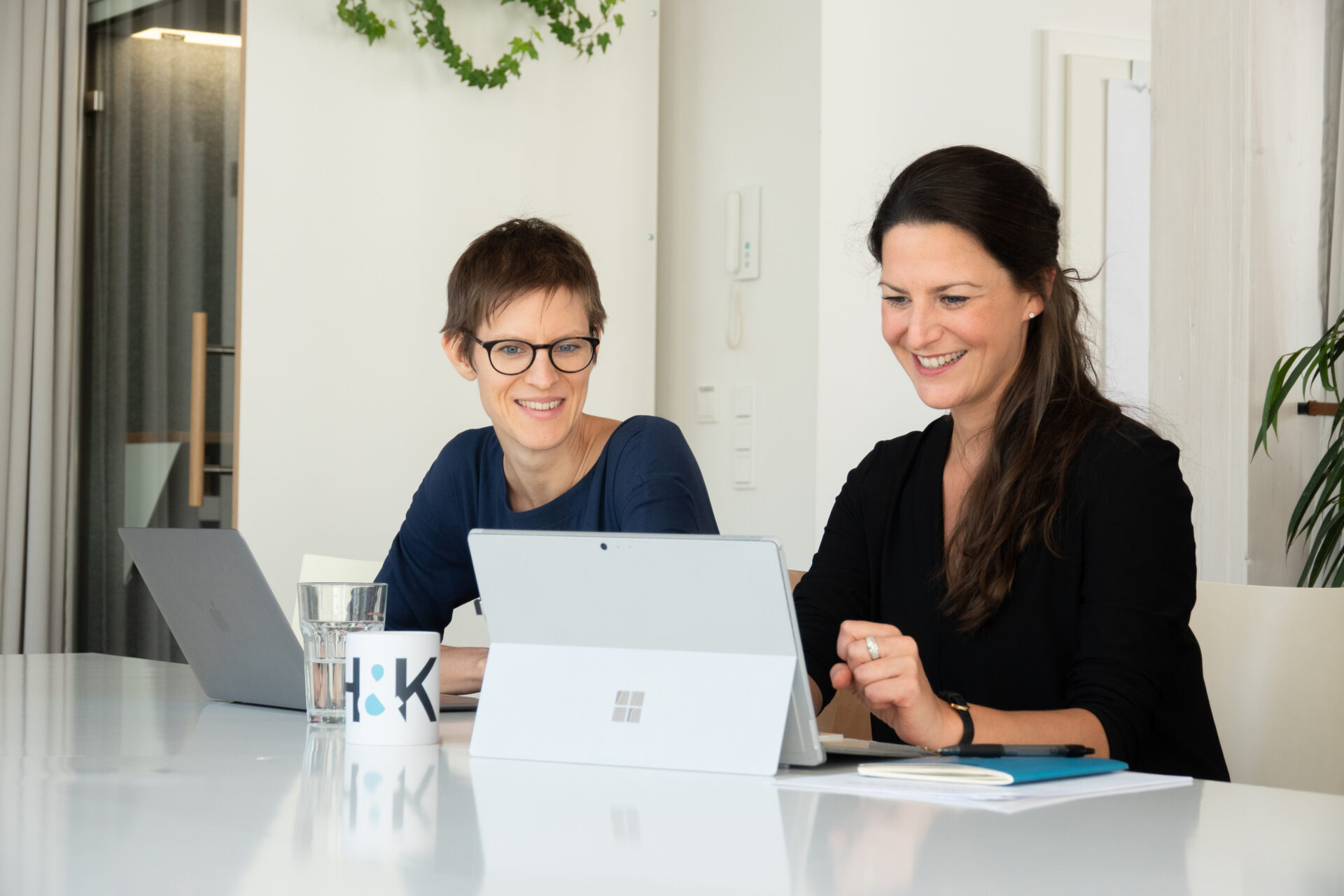Zwei Personen sitzen an einem Tisch mit Laptops und lächeln. Auf dem Tisch stehen eine Tasse und ein Glas Wasser. Im Hintergrund sind Pflanzen und ein Fenster zu sehen.