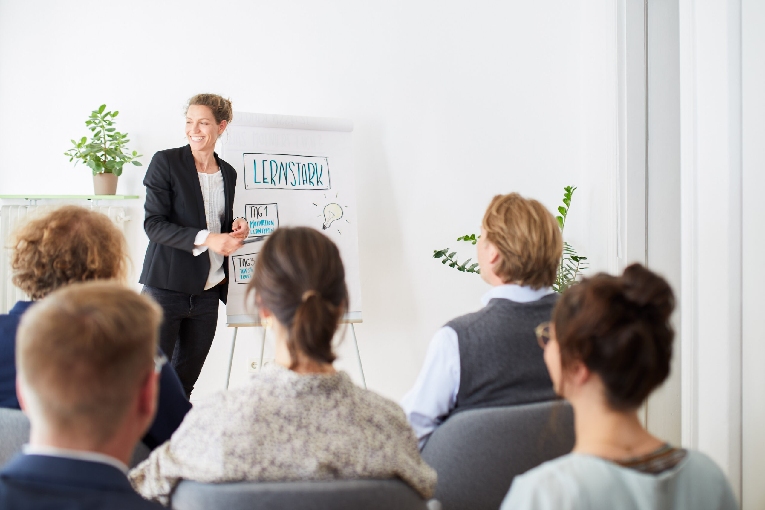 Eine Person steht lächelnd vor einem Flipchart mit der Aufschrift "LERNSTARK!" und spricht zu mehreren sitzenden Menschen in einem Raum mit Pflanzen.